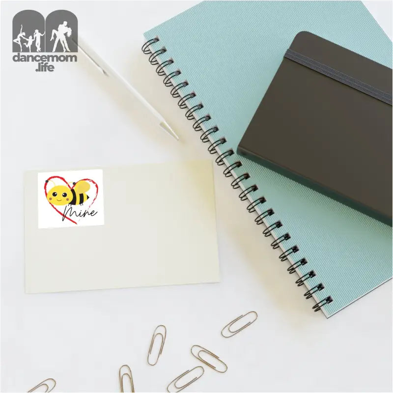 Spiral notebook with a mint green cover alongside paper clips and a small bee sticker.